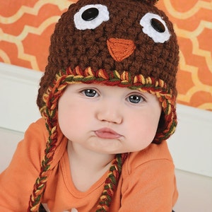May include: A baby wearing a brown crocheted turkey hat with a pom-pom on top. The hat has white eyes, a brown beak, and a red and yellow braided trim. The baby is wearing an orange long-sleeved shirt.