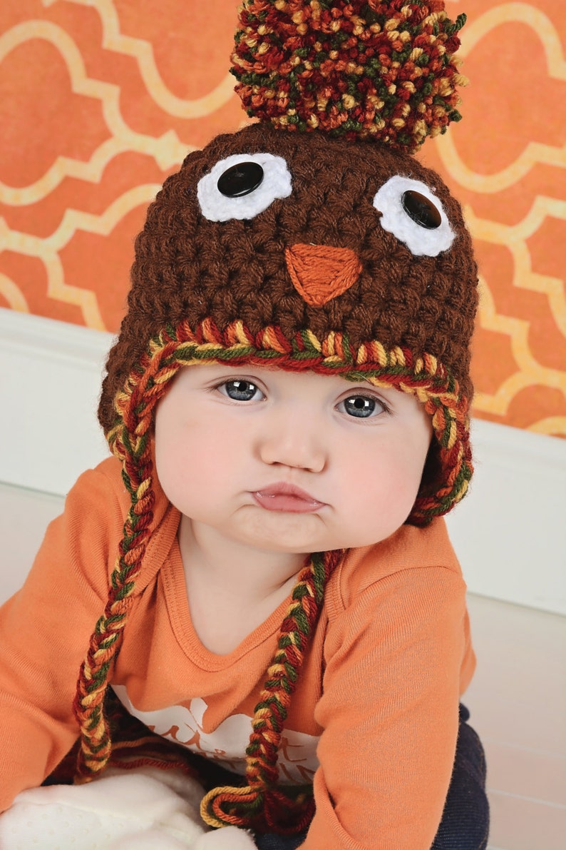May include: A baby wearing a brown crocheted turkey hat with a pom-pom on top. The hat has white eyes, a brown beak, and a red and yellow braided trim. The baby is wearing an orange long-sleeved shirt.