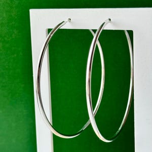 May include: A pair of large, polished silver-tone hoop earrings with a smooth, circular design. The earrings feature a thin, rounded metal wire construction and are suspended from the top edge of a white rectangular display frame.