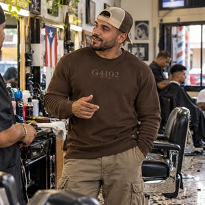May include: A dark brown long-sleeve crewneck sweatshirt featuring the text G4102 printed in a tonal brown shade on the chest. The sweatshirt is paired with tan cargo pants and a brown and beige mesh-back baseball cap.