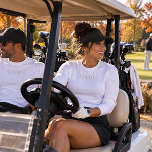 May include: Two individuals in a golf cart wearing matching white long-sleeve shirts with the text G4102 printed on the chest. The attire includes black baseball caps, a black visor, black pants, and a black skirt, with golf clubs stored in the back.