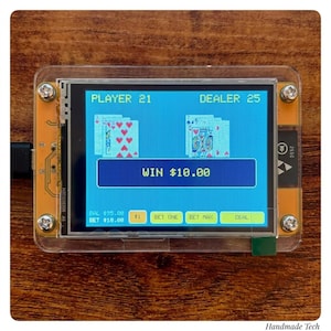 May include: A handheld electronic blackjack game device with a clear acrylic case and four corner screws. The screen displays a card game with the text PLAYER 21, DEALER 25, and WIN $10.00, along with buttons labeled BET ONE, BET MAX, and DEAL.