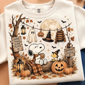 May include: A cream-colored crewneck sweatshirt featuring a graphic print of Snoopy and Woodstock in an autumn setting. The design includes a full moon, hanging laundry, pumpkins, lanterns, and wooden signs that read Good Things Scare People and Spooky Season.