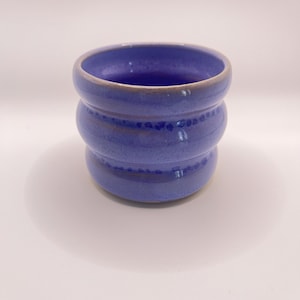 Op de afbeelding: Een kleine, cilindervormige keramische beker met een levendig maagdenblauw glazuur. De buitenkant heeft twee duidelijke horizontale ribbels rondom het lichaam, met subtiele donkerblauwe spikkels langs de verdiepte lijnen van de ribbels.