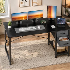 May include: A black computer desk with a wood grain finish, featuring a metal frame, two grey fabric drawers with black handles, and an open storage shelf. The desk surface holds a white keyboard, a mouse, a tablet, and three computer monitors.