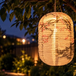 May include: A cylindrical paper lantern hanging from a tree branch, featuring a warm internal glow and a printed design of dark tree branches with delicate pink cherry blossoms. The lantern has a ribbed texture and is illuminated from within.