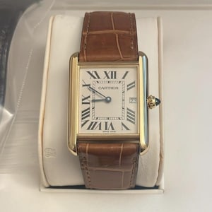 May include: A rectangular gold-tone wristwatch featuring a white dial with black Roman numeral hour markers, a date window at the three o'clock position, and the Cartier logo. The watch has a brown leather strap with a textured crocodile-like pattern.