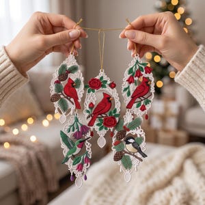 May include: A set of five white lace ornaments featuring embroidered birds and botanical elements. The designs include red cardinals, a hummingbird, and a chickadee, accented with pinecones, red roses, and green foliage, all suspended by gold strings.