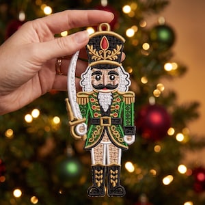 May include: An embroidered nutcracker ornament featuring a green jacket with gold trim, a black hat with a red accent, and a white sword. The figure has a black mustache, white hair, and black boots, rendered in detailed thread work with a hanging loop.