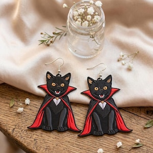 May include: A pair of embroidered dangle earrings featuring black cats dressed in red-lined capes with white collars. The cats have yellow eyes and visible white fangs. Each earring is attached to a silver-toned metal hook for pierced ears.