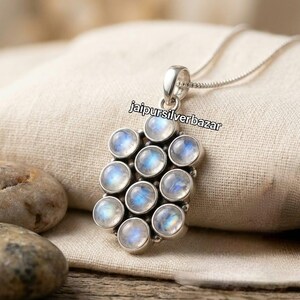 May include: A silver pendant necklace featuring ten round, cabochon-cut moonstones arranged in a cluster. The stones exhibit a blue adularescence. The pendant is attached to a thin silver chain. The text jaipursilverbazar is visible on the image.