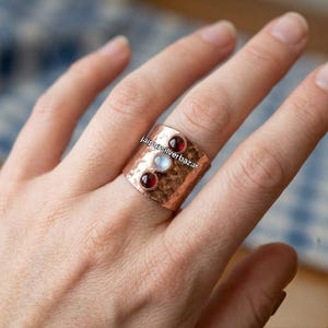 May include: A wide hammered copper band ring featuring three vertically aligned cabochon gemstones: a deep red garnet at the top, a translucent white moonstone in the centre, and a deep red garnet at the bottom, with the text jaipursilverbazar.