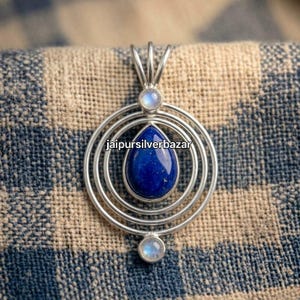 Op de afbeelding: Een zilveren hanger met een centrale druppelvormige lapis lazuli steen omringd door drie concentrische zilveren cirkels, geaccentueerd met twee kleine ronde maanstenen aan de boven- en onderkant, met de tekst jaipursilverbazar.