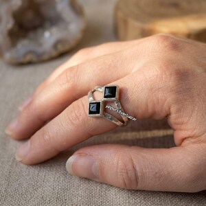 Puede incluir: Un anillo de plata con dos piedras negras de corte cuadrado engastadas en una banda de estilo bypass. El metal tiene un acabado pulido y el diseño incluye un vástago dividido que sostiene las piedras en una disposición diagonal. El texto jaipursilverbazar es visible.