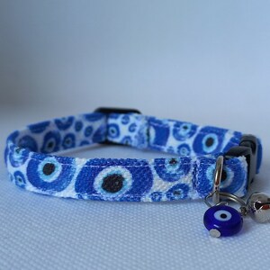 May include: A white cat collar with a blue and white evil eye pattern. The collar has a silver bell and a charm with a blue evil eye.