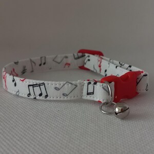 Peut inclure: Collier pour chat blanc avec une boucle rouge et une clochette argentée. Le collier est décoré d'un motif de notes de musique en noir et gris.