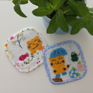 May include: Two square fabric coasters with embroidered designs. One coaster features a yellow cat with a fish, flowers, and a branch. The other coaster features a yellow cat with an umbrella, a frog, and flowers.