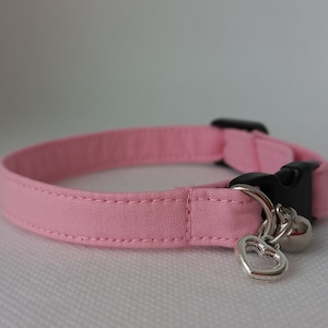 Pode incluir: Um colar de gato de tecido rosa com uma fivela preta e um pingente de coração prateado com um sino.