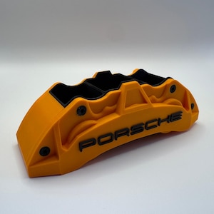 May include: A bright orange automotive brake caliper model featuring the black text PORSCHE embossed on the front. The object has a matte finish with black interior detailing and small black circular accents along the outer edges of the structure.