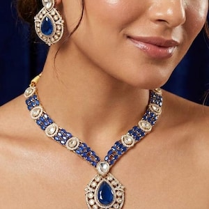 May include: A gold-toned necklace and matching earring set featuring deep blue, pear-shaped central stones surrounded by smaller, clear, round-cut stones. The necklace has a multi-layered chain design with blue and clear stones in a metal setting.