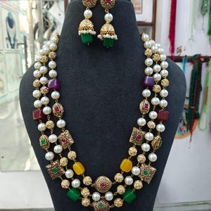 Op de afbeelding: Een drie-rijige ketting en bijpassende jhumka-oorbellen met witte parels, goudkleurige metalen tussenstukjes en veelkleurige vierkante en ovale kralen in rood, paars, geel en groen. De oorbellen hebben een knopje en een klokvormige hanger.