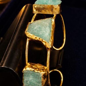 Può includere: Un bracciale rigido color oro con tre pietre grezze color blu-verde chiaro, incastonate in montature metalliche testurizzate e irregolari. Il design aperto è composto da sottili fasce metalliche curve che collegano le pietre, creando un aspetto strutturato.
