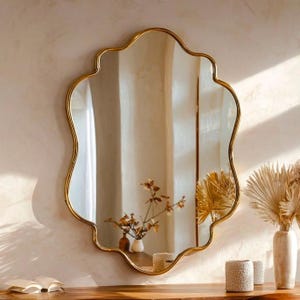 May include: A wall-mounted mirror featuring a scalloped, wavy gold frame. The reflective glass surface displays a clear view of a room interior, including a small vase with dried branches and a portion of a white wall with soft lighting.