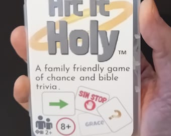 以下が含まれることがあります： Hit It Holyカードゲームが入った透明なプラスチックケース。ラベルには黄色の光輪が付いた灰色の文字でタイトルが記され、サブタイトル「A family friendly game of chance and bible trivia」、2人以上のプレイヤーと8歳以上を示すアイコンが表示されています。