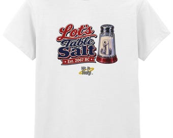 以下が含まれることがあります： 白い半袖Tシャツで、中に人物が入った塩入れのグラフィックが描かれています。テキストには「Lot's Table Salt Est. 2067 BC」および「Hit It Holy」と記されています。デザインは赤、青、黄の色を使用しています。