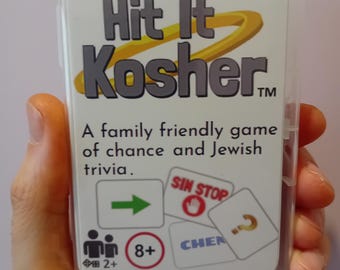 以下が含まれることがあります： Hit It Kosherというタイトルのカードゲームが入った透明なプラスチックケース。白いラベルには、黄色のリングが付いた灰色の文字でタイトルが記され、サブタイトル「A family friendly game of chance and Jewish trivia」、2人以上のプレイヤーと8歳以上を示すアイコンが印刷されています。