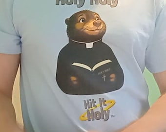 以下が含まれることがあります： 聖職者の服を着た茶色のモグラが「Holy Bible」と書かれた開いた本を持っているグラフィックが描かれた水色のTシャツ。モグラの上には「Holy Moly」、下には黄色の光輪のグラフィックと共に「Hit It Holy」というテキストがプリントされています。