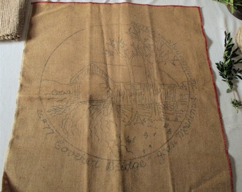 May include: A brown burlap fabric with a red border, featuring a black and white drawing of a covered bridge with trees and the text "The Covered Bridge of Thompsons 1877" in a circle.
