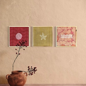 Op de afbeelding: Drie vierkante ingelijste kunstwerken met verschillende patronen: een dieprode print met een witte schelp, een mosterdgele gestreepte print met een witte zeester en een roze bloemenprint met een witte golvende lijn in het midden.