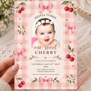 Op de afbeelding: Een rechthoekige verjaardagsuitnodiging met een roze en wit gingham-achtergrond, versierd met rode kersen, witte bloesems en roze strikken. Het midden toont een foto van een baby met de tekst Olivia Turns One Sweet Cherry.