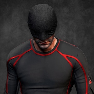 Pode incluir: Uma peça preta texturizada para a cabeça que oculta os olhos e uma camisola preta de manga comprida com detalhes de debrum vermelho ao longo dos ombros, peito e braços. O tecido parece liso e justo, com um detalhe vermelho na manga esquerda.