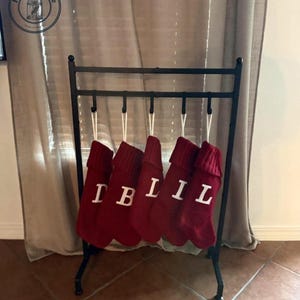 May include: A black metal stand with five hooks holding five red knit Christmas stockings. Each stocking features a white letter, spelling out the name LILIB, with a ribbed cuff at the top and a white hanging loop attached to each hook.