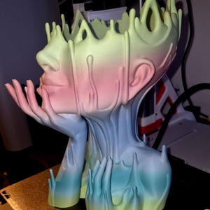May include: A 3D printed planter shaped like a human head with a crown-like top edge. The surface features a smooth gradient transition from yellow at the top to pink in the middle and blue at the base. Hands are sculpted supporting the chin.