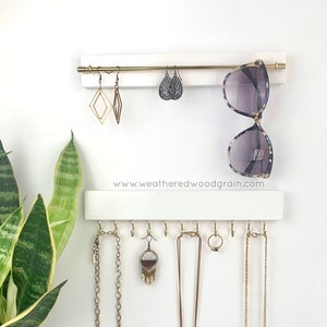 Könnte beinhalten: Ein weißer Wand-Schmuckorganizer mit zwei horizontalen Stangen. Die obere Stange ist goldfarben für Ohrringe und Sonnenbrillen. Die untere Stange verfügt über goldene Haken für Halsketten und Ringe. Der Text www.weatheredwoodgrain.com ist in der Mitte aufgedruckt.