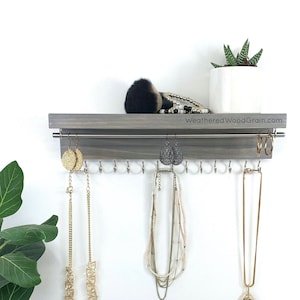 May include: A gray wooden jewelry organizer with hooks for necklaces and earrings. The organizer has a shelf on top for storing other items. The organizer is mounted on a white wall. The text "WeatheredWoodGrain.com" is printed on the organizer.