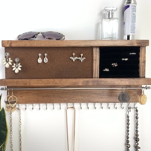 May include: A wooden jewelry organizer with a corkboard section for earrings, a velvet-lined compartment for rings, and a row of hooks for necklaces. The organizer is mounted on a wall and has a dark brown stain.