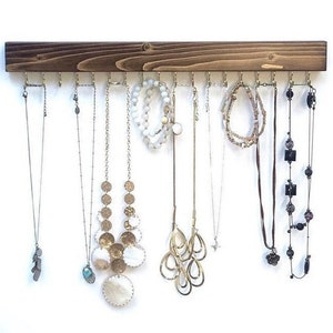 Pu&ograve; includere: Un organizer da parete per gioielli in legno tinto marrone scuro, caratterizzato da una tavola orizzontale con molteplici ganci in metallo dorato. Diverse collane, bracciali e catenine di varie lunghezze e stili sono appesi ai ganci.