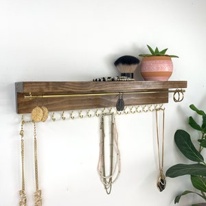 May include: A wooden shelf with a gold metal rod for hanging necklaces and earrings. The shelf has a dark brown stain and is mounted on a white wall. There are several necklaces and earrings hanging from the shelf, as well as a small potted plant.