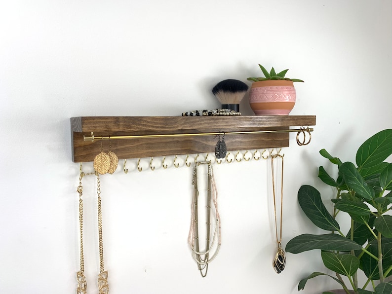 May include: A wooden shelf with a gold metal rod for hanging necklaces and earrings. The shelf has a dark brown stain and is mounted on a white wall. There are several necklaces and earrings hanging from the shelf, as well as a small potted plant.
