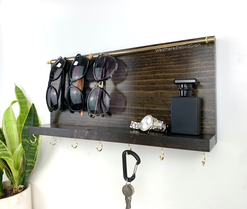May include: A dark brown wooden wall mounted organiser with a shelf, hooks, and a rod for hanging items. The organiser has a gold metal rod for hanging sunglasses, a shelf for a watch and a bottle of cologne, and five gold hooks for hanging keys. The text "Weathered WoodGrain" is printed on the organiser.