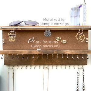 May include: A wooden jewelry organizer with two shelves. The top shelf has a corkboard for stud earrings and a metal rod for dangle earrings. The bottom shelf has hooks for necklaces. The organizer is 8 inches tall and the shelves are 2 inches deep.