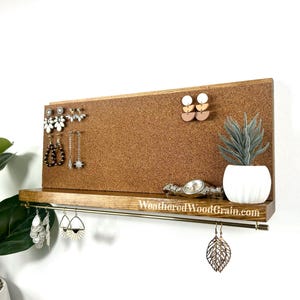 May include: A wooden jewelry organizer with a corkboard backing and a shelf. The shelf holds a watch and a small potted plant. Various earrings are displayed on the board and hanging from the shelf. The website address "WeatheredWoodGrain.com" is visible.