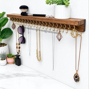 May include: A dark brown wooden wall-mounted shelf featuring a row of gold-toned metal hooks along the bottom edge for hanging jewelry. The shelf displays a makeup brush, a small white potted succulent, and various necklaces and sunglasses.