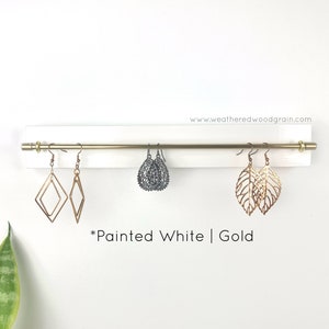May include: A white painted wooden wall-mounted earring organiser featuring a horizontal gold metal rod. The display holds three pairs of earrings: gold geometric diamonds, dark grey filigree teardrops, and gold leaf-shaped cutouts. Text reads www.weatheredwoodgrain.com and *Painted White | Gold.