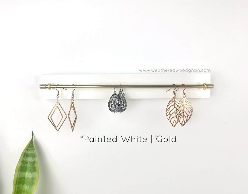 May include: A white painted wooden wall-mounted earring organiser featuring a horizontal gold metal rod. The display holds three pairs of earrings: gold geometric diamonds, dark grey filigree teardrops, and gold leaf-shaped cutouts. Text reads www.weatheredwoodgrain.com and *Painted White | Gold.