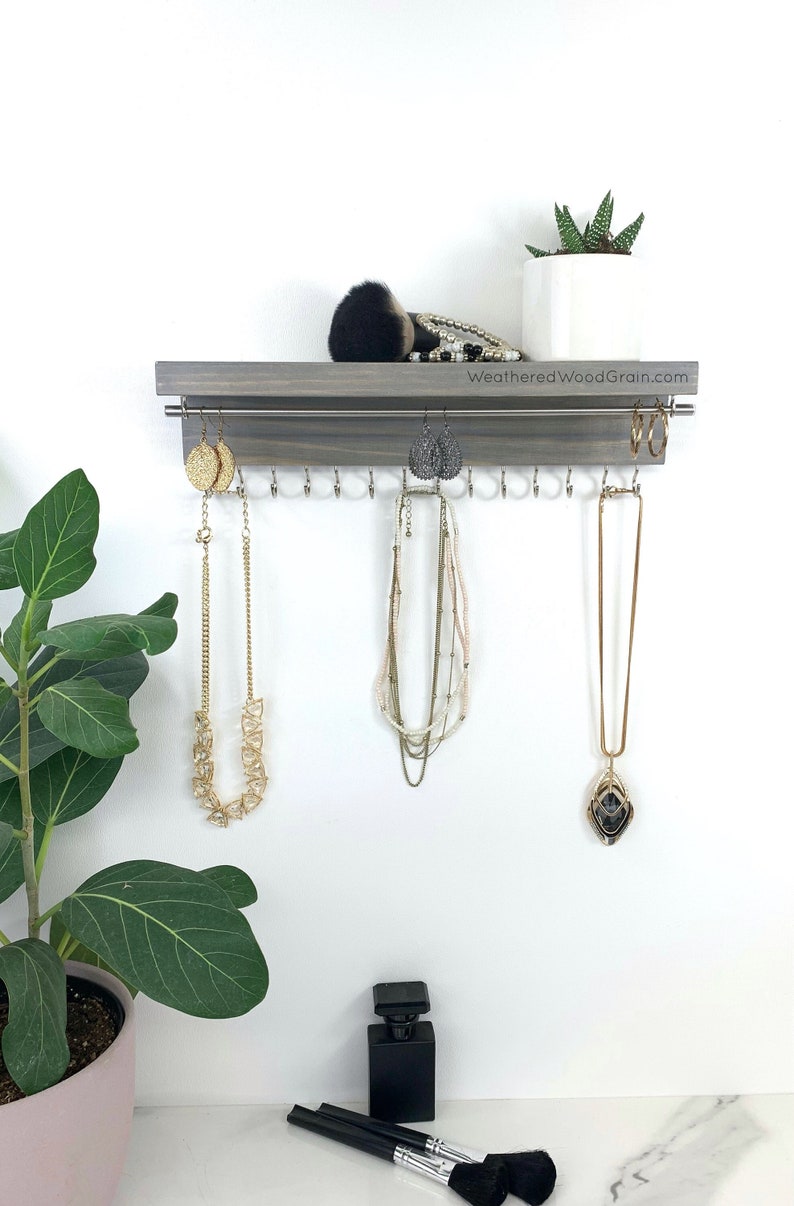 May include: A gray wooden jewelry organizer with hooks for necklaces and earrings. The organizer has a shelf on top for storing other items. The organizer is mounted on a white wall. The text "WeatheredWoodGrain.com" is printed on the organizer.